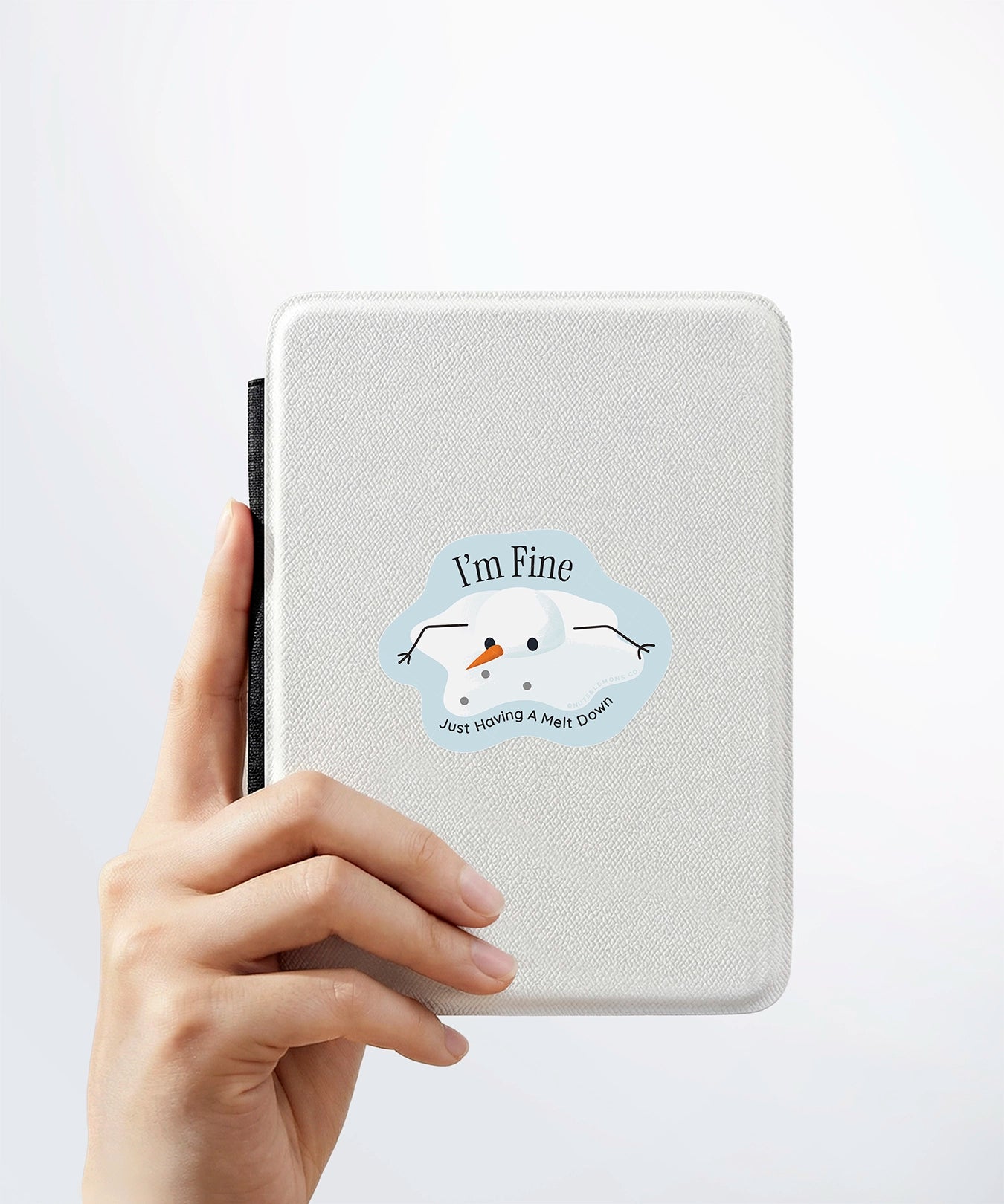 A hand holding a white kindle cover with a Nuts & Lemons Co. diecut sticker on it featuring a melted snowman  with the phrase “I’m Fine, Just Having a Meltdown”