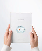 A pair of hands holding a white notebook with a Nuts & Lemons Co. diecut sticker on it featuring a melted snowman  with the phrase “I’m Fine, Just Having a Meltdown”