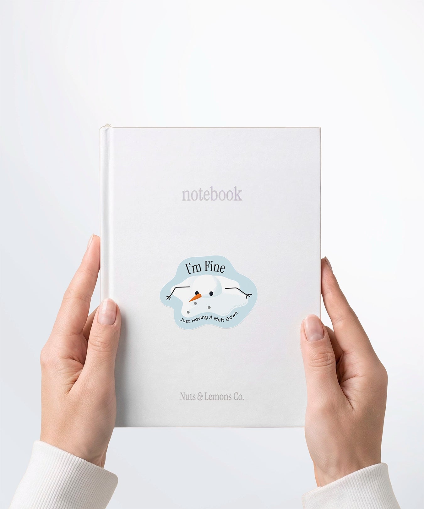 A pair of hands holding a white notebook with a Nuts & Lemons Co. diecut sticker on it featuring a melted snowman  with the phrase “I’m Fine, Just Having a Meltdown”