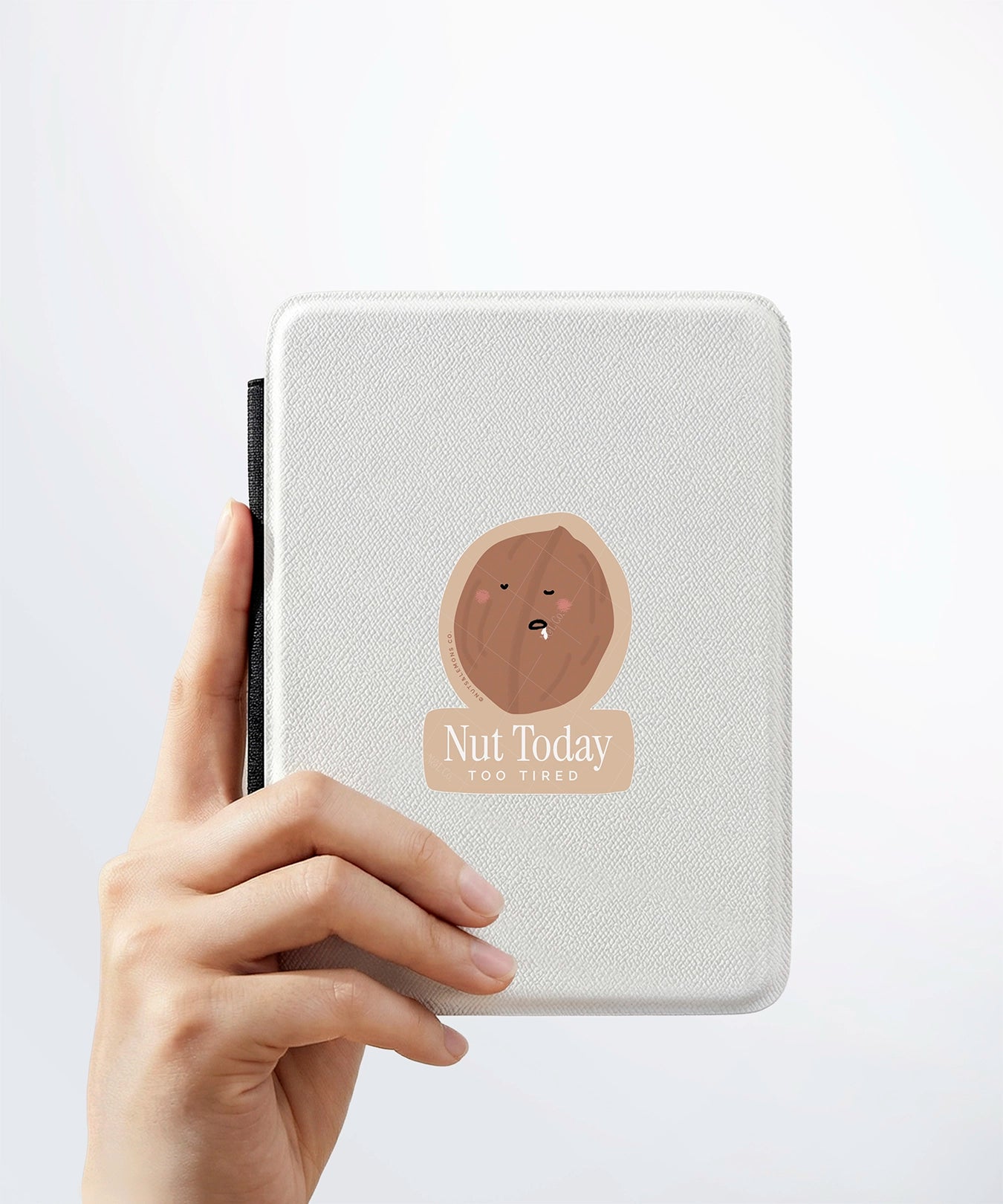 A hand holding a white kindle cover with a Nuts & Lemons Co. diecut sticker on it featuring a walnut drooling asleep with the phrase “Nut today too tired”