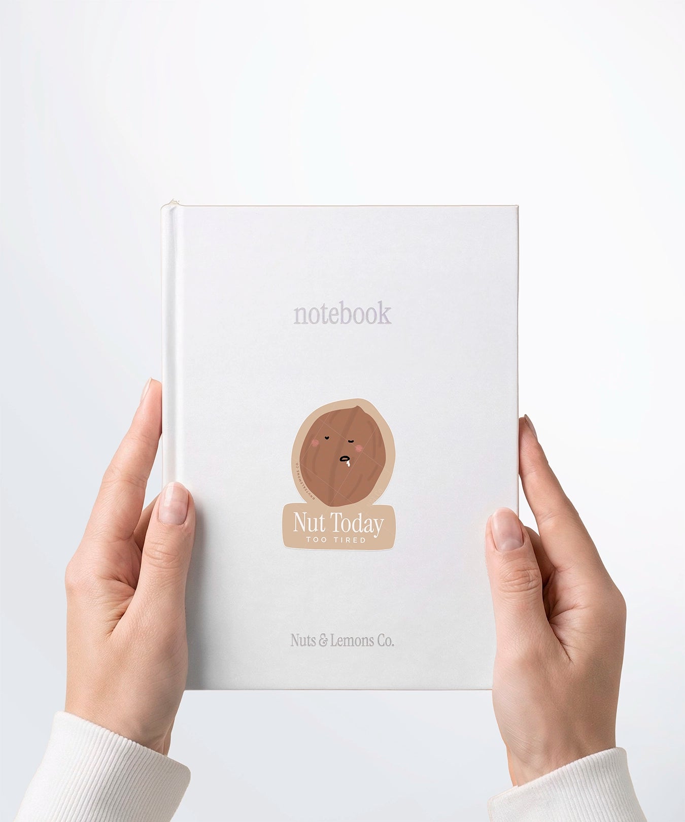 A pair of hands holding a white notebook with a Nuts & Lemons Co. diecut sticker on it featuring a walnut drooling asleep with the phrase “Nut today too tired”