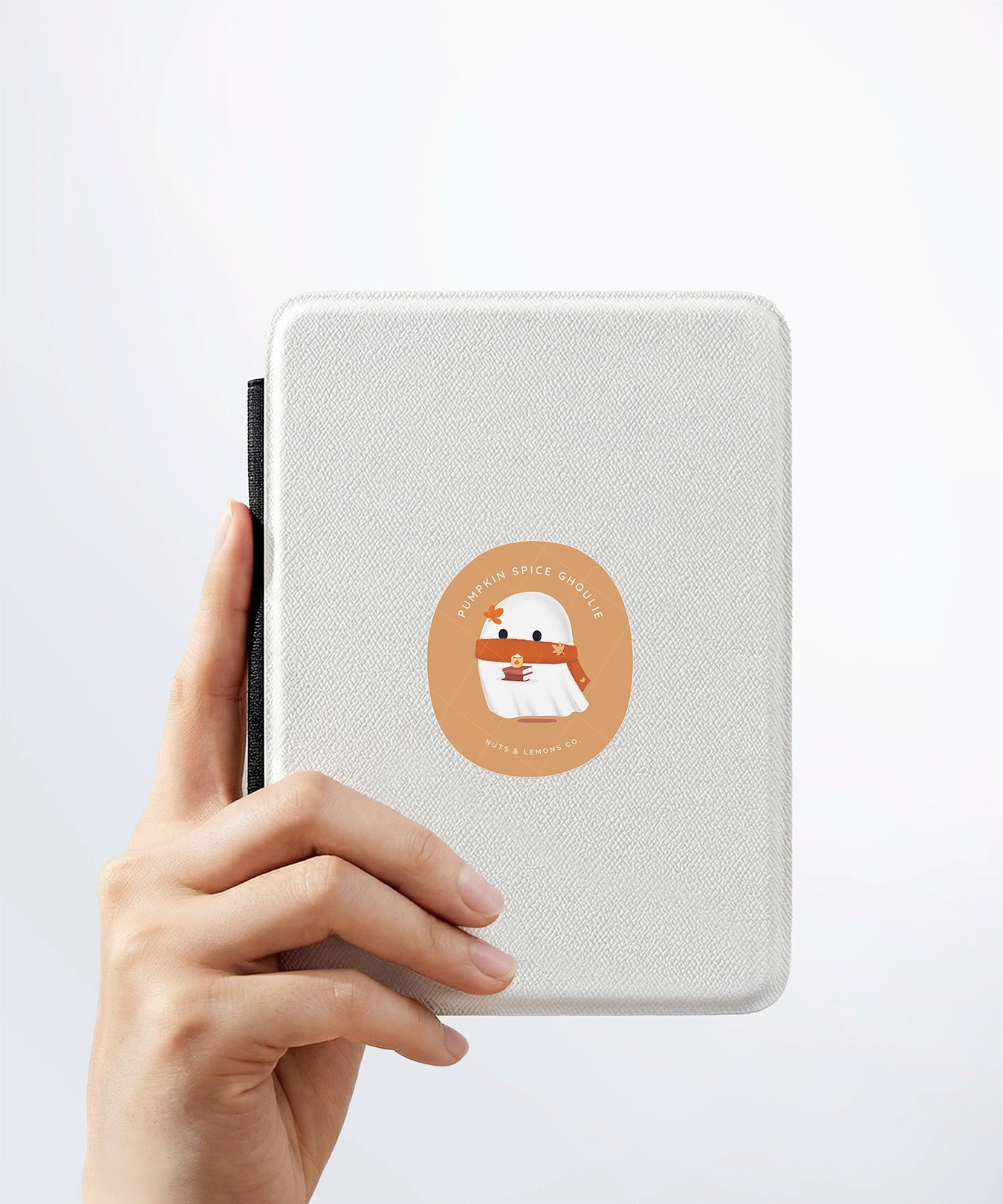 A hand holding a white kindle cover with a Nuts & Lemons Co. diecut sticker on it featuring a cute ghostie with autumn books, scarf and pumpkin spice latte with the phrase “Pumpkin Spice Ghoulie”
