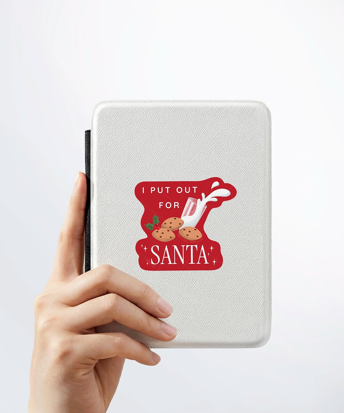 A hand holding a white kindle cover with a Nuts & Lemons Co. diecut sticker on it featuring a bunch of cookies and spilling milk with the phrase “Put Out for Santa”