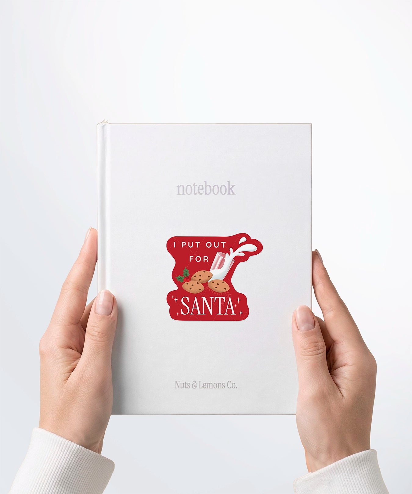 A pair of hands holding a white notebook with a Nuts & Lemons Co. diecut sticker on it featuring a bunch of cookies and spilling milk with the phrase “Put Out for Santa”