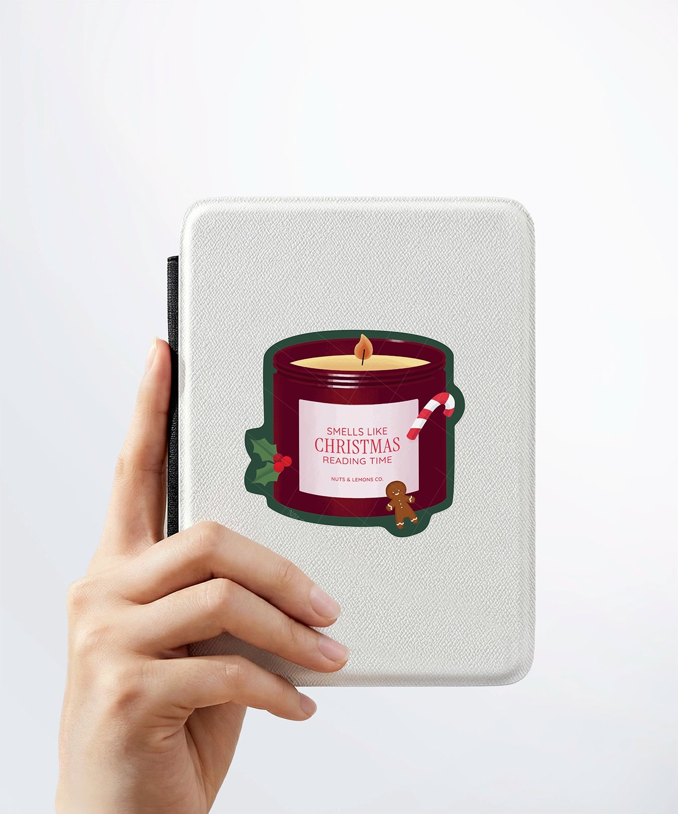 A hand holding a white kindle cover with a Nuts & Lemons Co. diecut sticker on it featuring a Christmas candle with candy cane, gingerbread and a label with the phrase “Smells Like Christmas Reading Time”