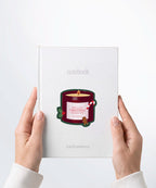 A pair of hands holding a white notebook with a Nuts & Lemons Co. diecut sticker on it featuring a Christmas candle with candy cane, gingerbread and a label with the phrase “Smells Like Christmas Reading Time”