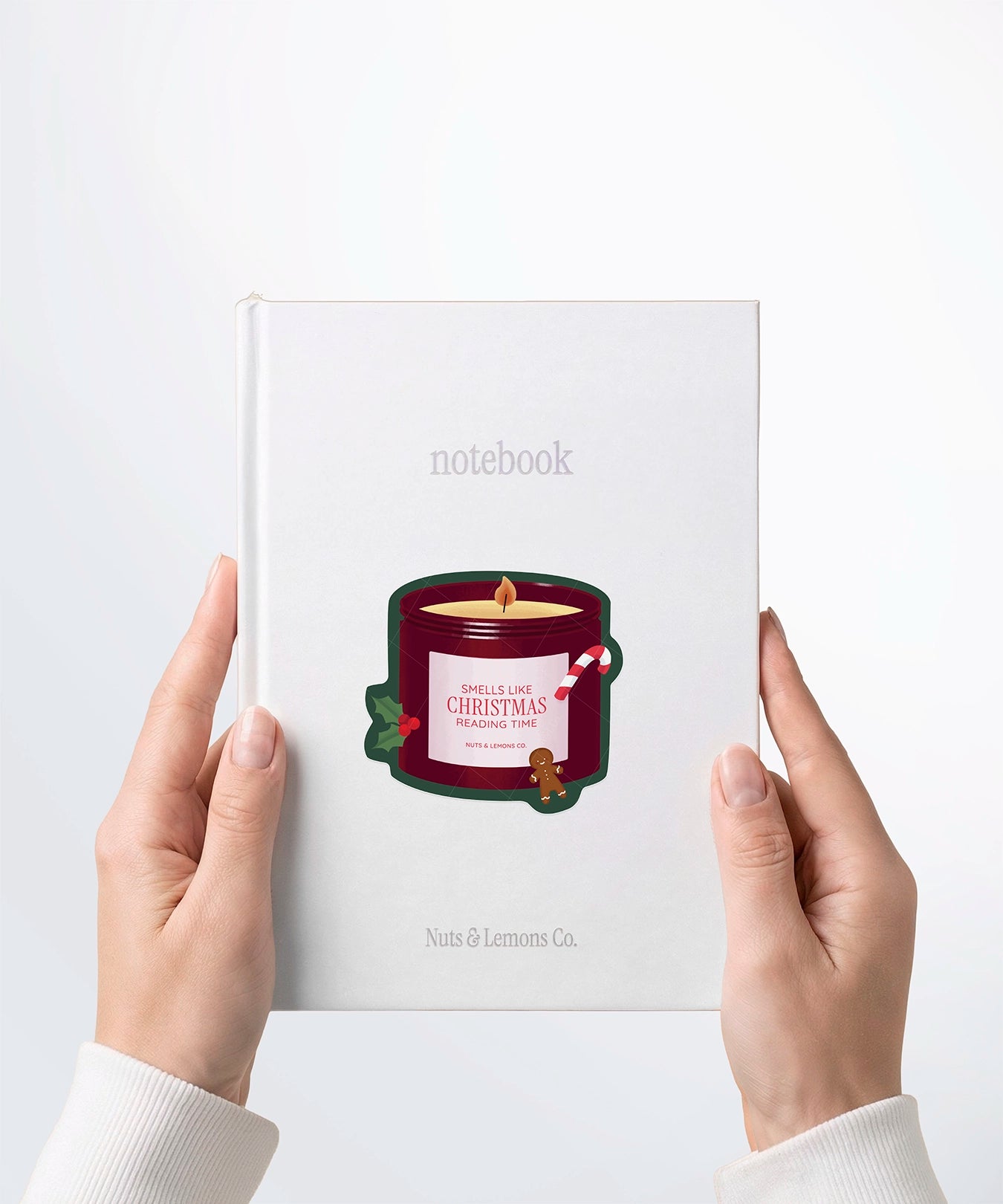A pair of hands holding a white notebook with a Nuts & Lemons Co. diecut sticker on it featuring a Christmas candle with candy cane, gingerbread and a label with the phrase “Smells Like Christmas Reading Time”