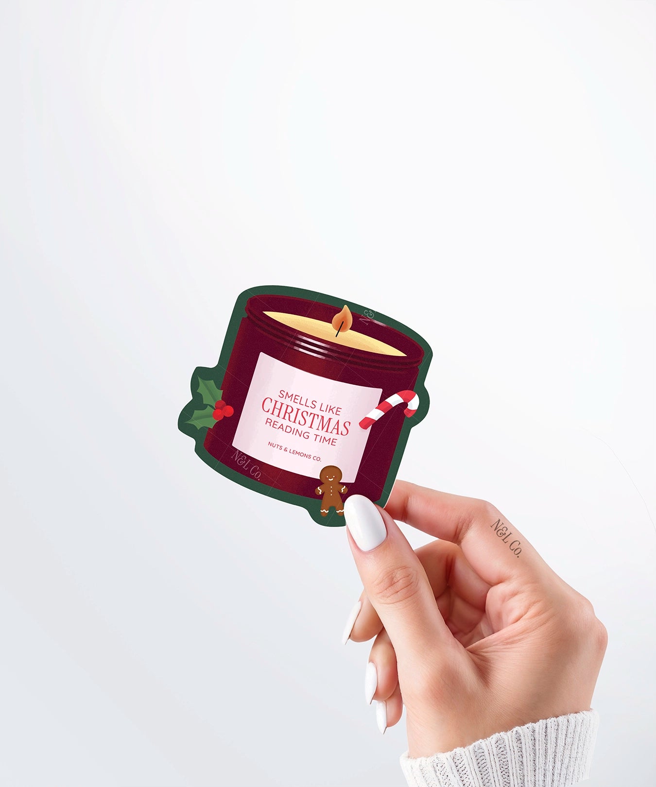 Hand holding a Nuts & Lemons Co. diecut sticker featuring a Christmas candle with candy cane, gingerbread and a label with the phrase “smells like Christmas reading time”