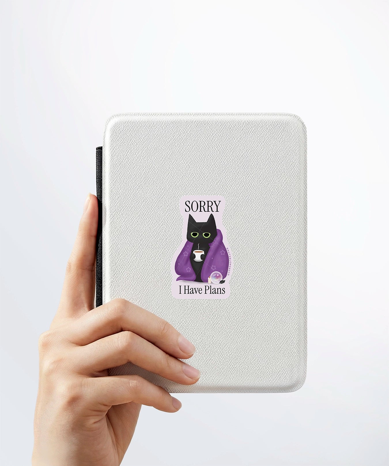 A hand holding a white kindle cover with a Nuts & Lemons Co. diecut sticker on it featuring a black cat in a starry blanket holding a mug and crystals with the phrase “Sorry I have Plans”