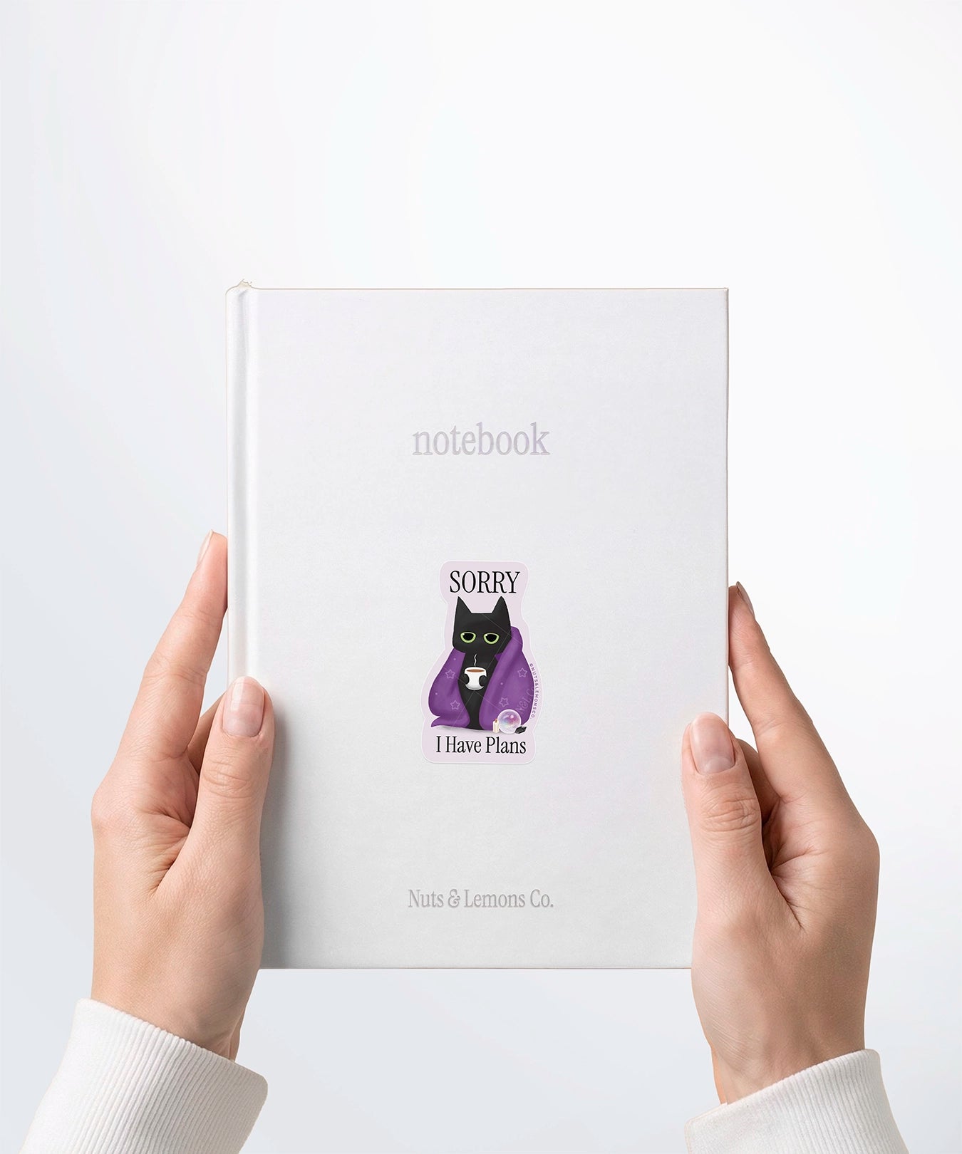 A pair of hands holding a white notebook with a Nuts & Lemons Co. diecut sticker on it featuring a black cat in a starry blanket holding a mug and crystals with the phrase “Sorry I have Plans”