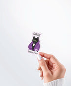 Hand holding a Nuts & Lemons Co. diecut sticker featuring a black cat in a starry blanket holding a mug and crystals with the phrase “Sorry I Have Plans”