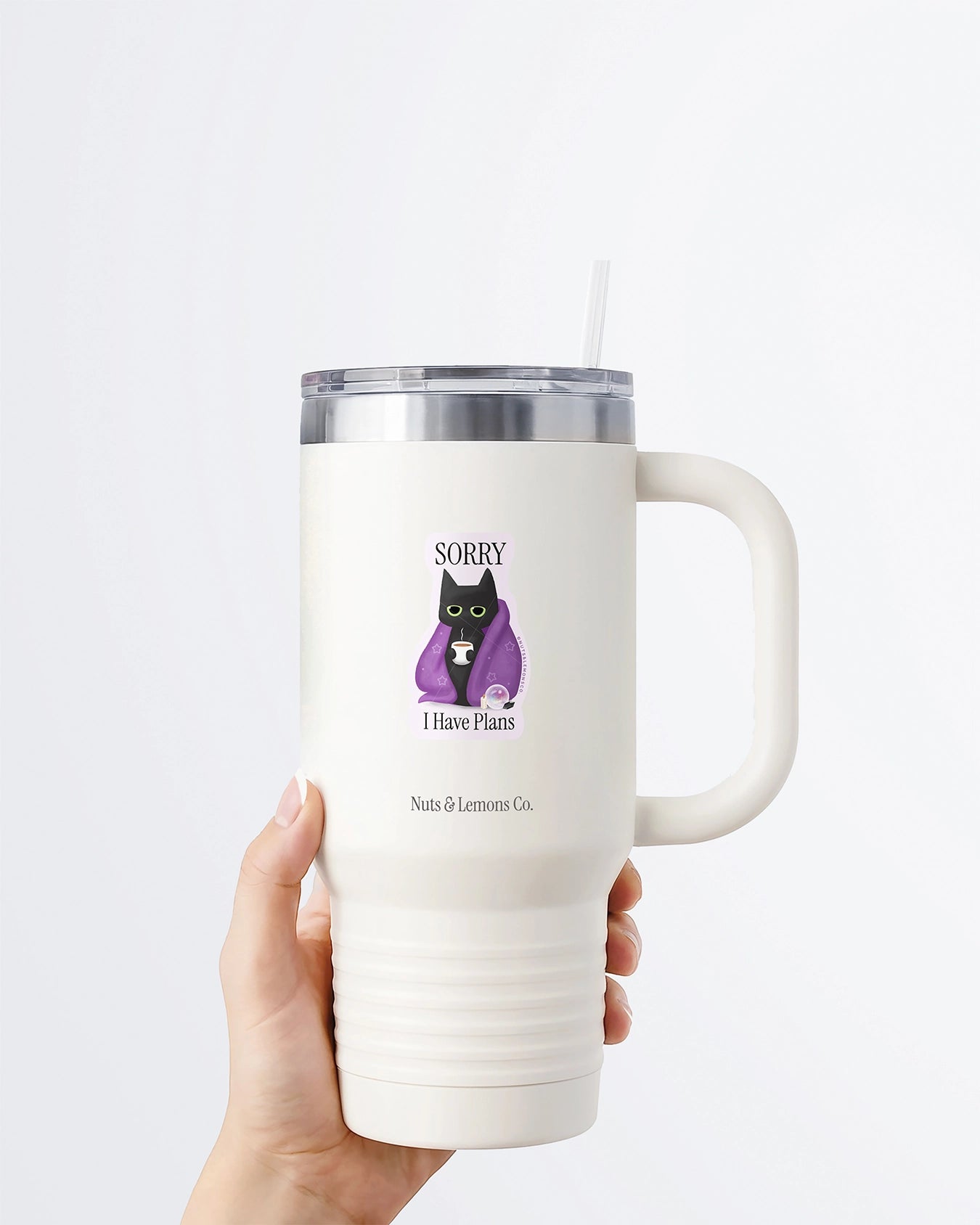 A hand holding a white tumbler with a Nuts & Lemons Co. diecut sticker on it featuring a black cat in a starry blanket holding a mug and crystals with the phrase “Sorry I Have Plans”