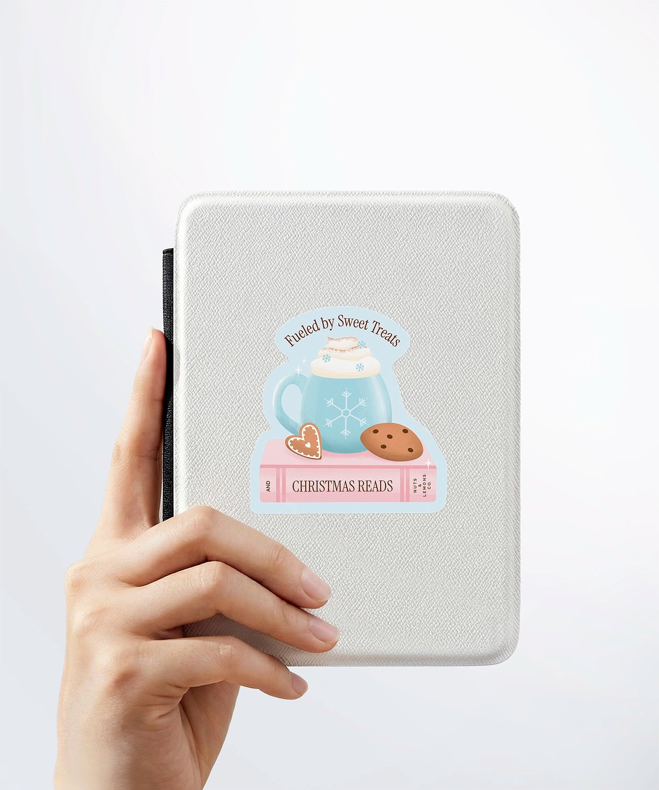A hand holding a white kindle cover with a Nuts & Lemons Co. diecut sticker on it featuring a blue snowflake mug on a pink book and cookies with the phrase “Fueled by Sweet Treats and Christmas Reads”