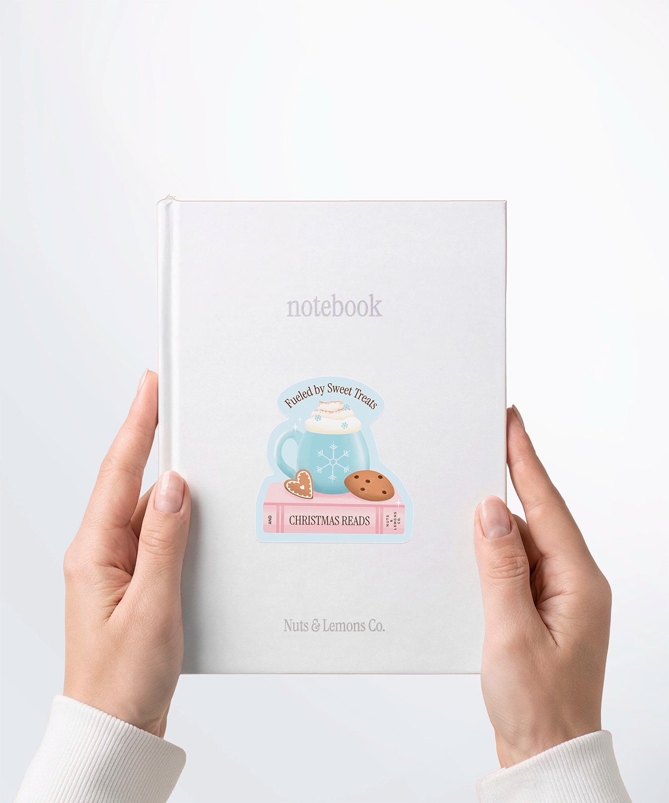 A pair of hands holding a white notebook with a Nuts & Lemons Co. diecut sticker on it featuring a blue snowflake mug on a pink book and cookies with the phrase “Fueled by Sweet Treats and Christmas Reads”
