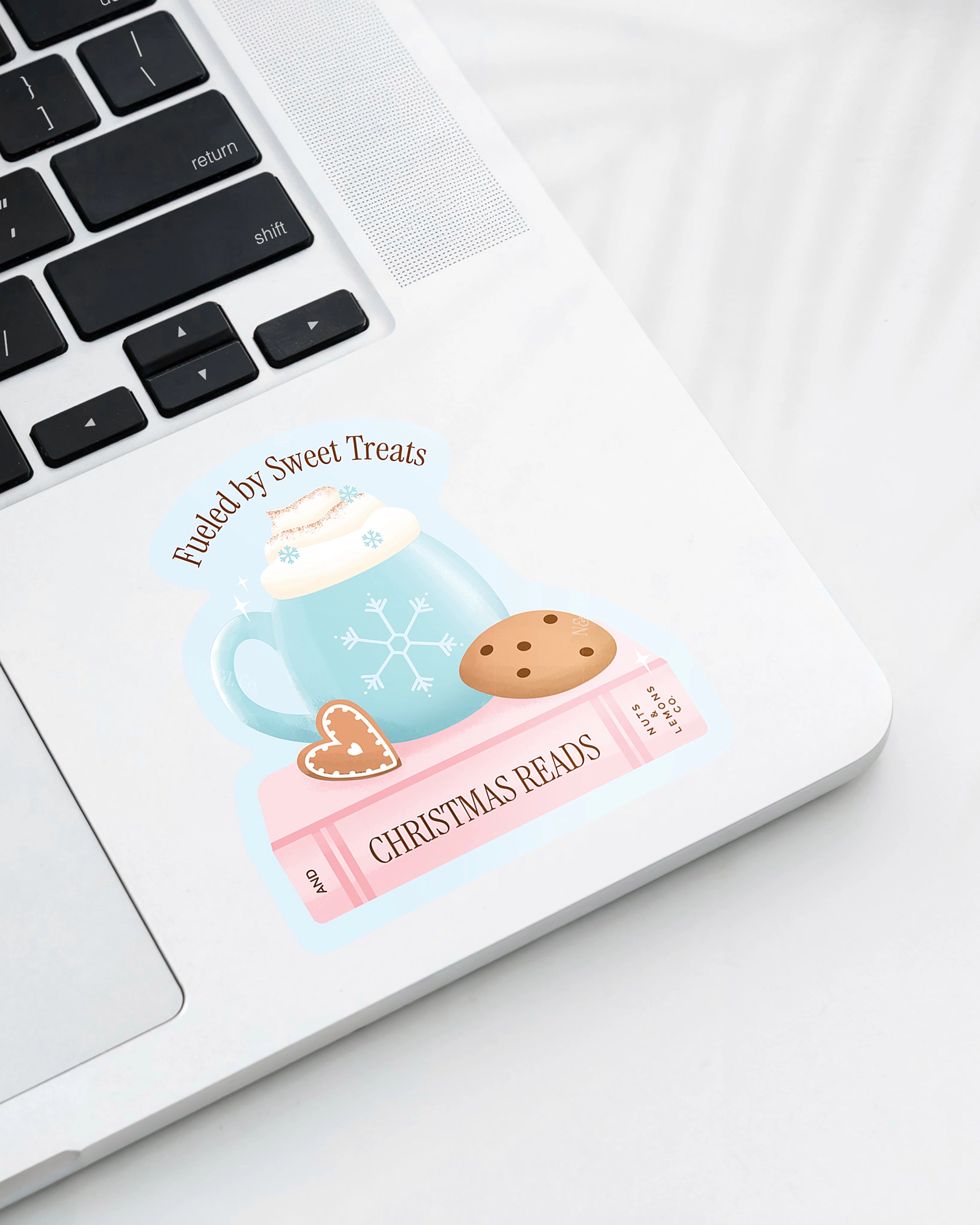 Laptop displaying a Nuts & Lemons Co. diecut sticker featuring a blue snowflake mug on a pink book and cookies with the phrase “fueled by sweet treats and Christmas reads”
