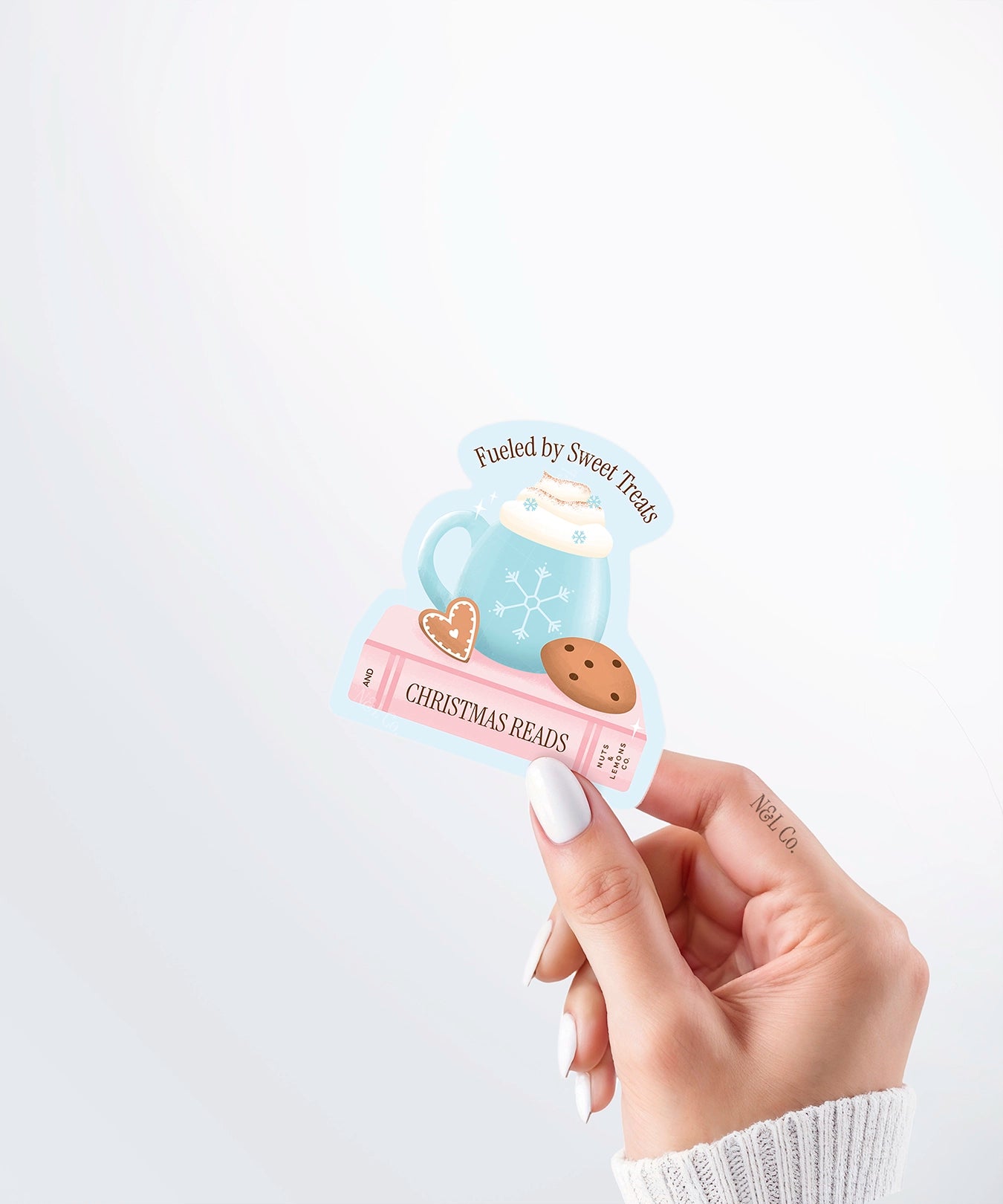Hand holding a Nuts & Lemons Co. diecut sticker featuring a a blue snowflake mug on a pink book and cookies with the phrase “fueled by sweet treats and Christmas reads”