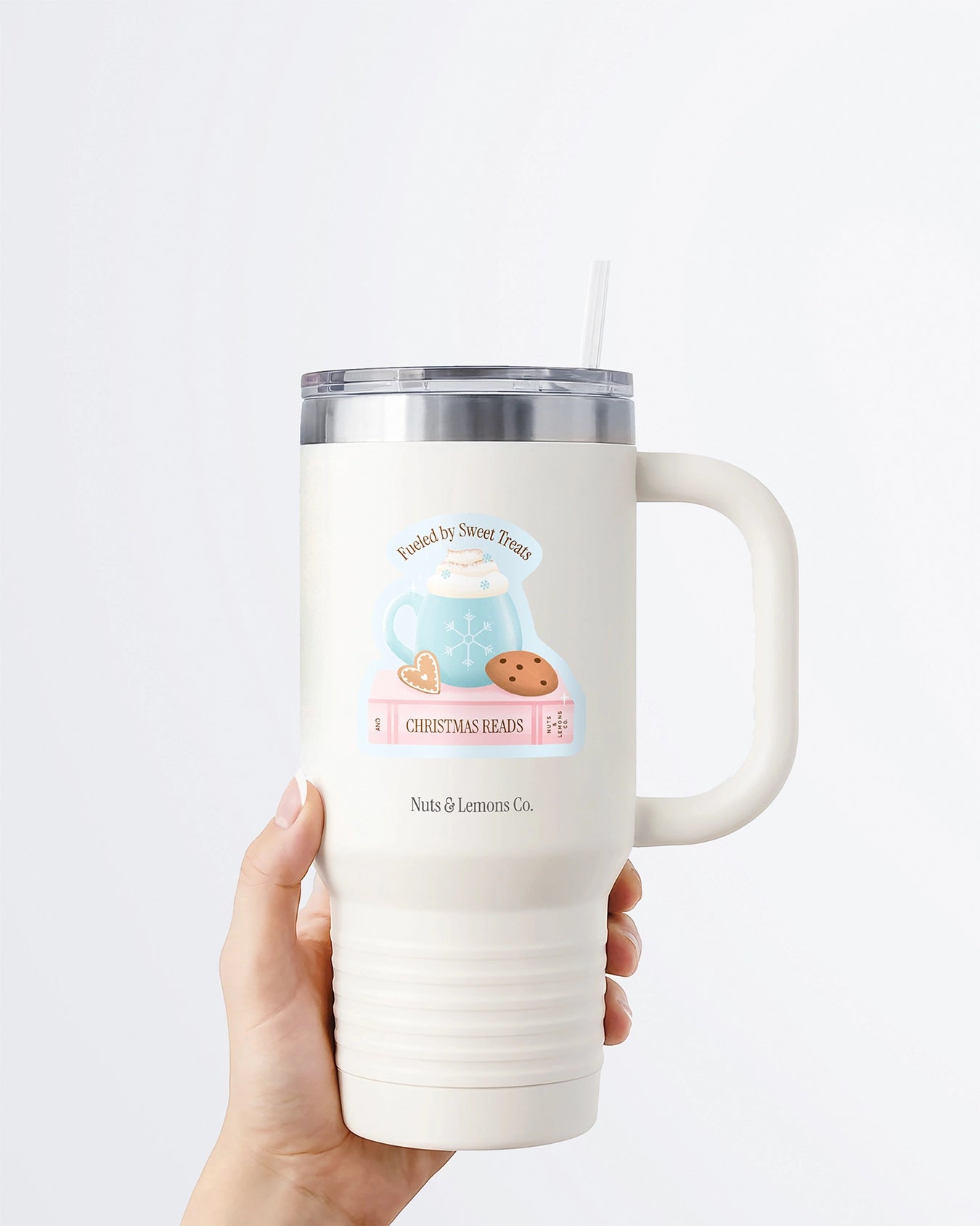 Hand holding a tumbler displaying Nuts & Lemons Co. diecut sticker featuring  a blue snowflake mug on a pink book and cookies with the phrase “fueled by sweet treats and Christmas reads”