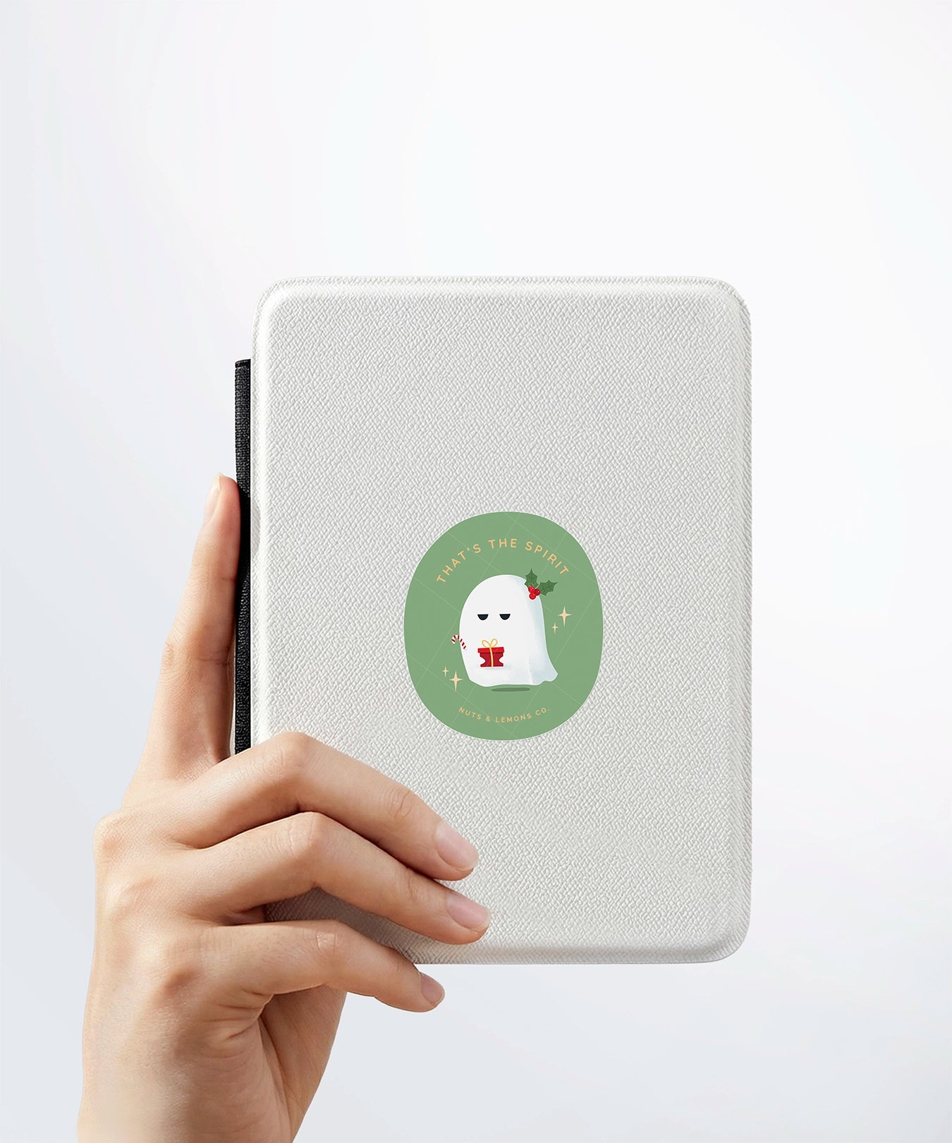 A hand holding a white kindle cover with a Nuts & Lemons Co. diecut sticker on it featuring a cute pouting and grumpy ghostie with a Christmas present with the phrase “That’s the Spirit”