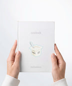 A pair of hands holding a white notebook with a Nuts & Lemons Co. diecut sticker on it featuring a yogurt cup with the phrase “Yogurt Has More Culture”