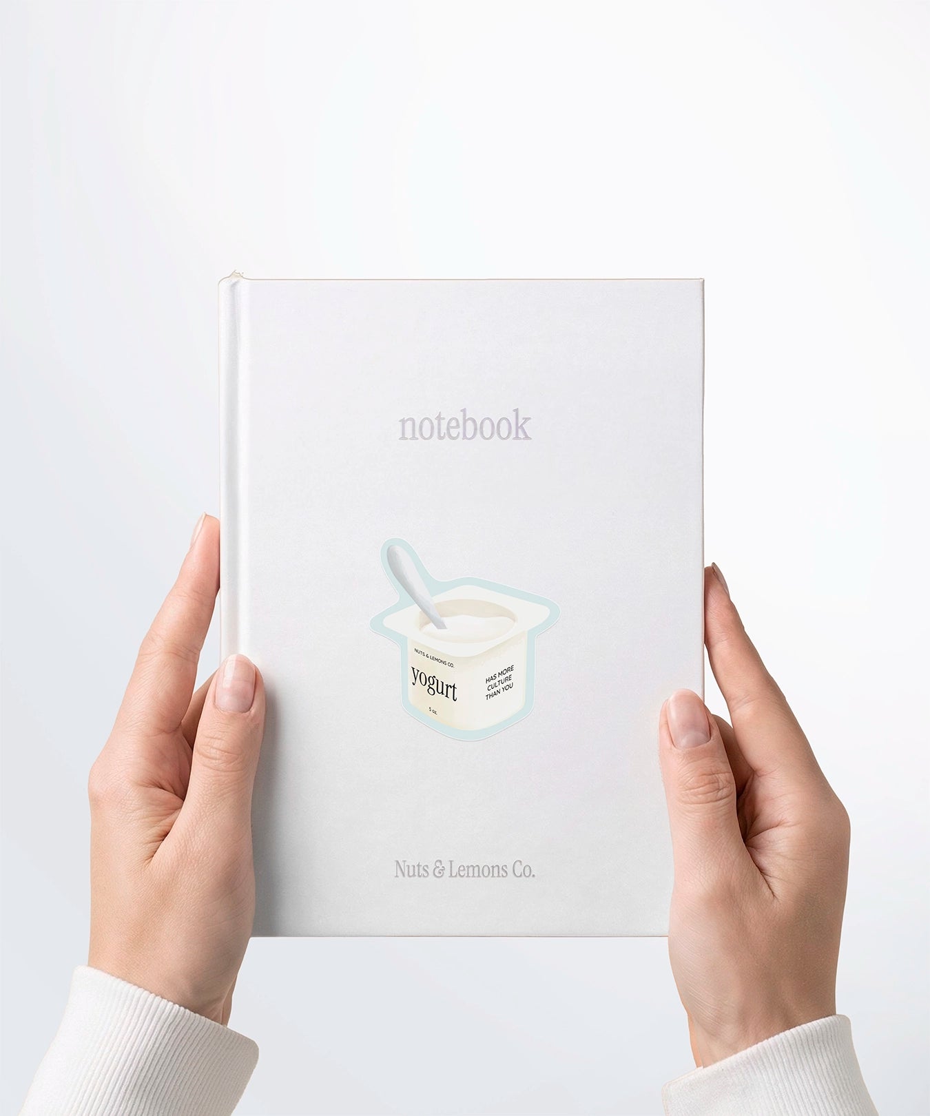 A pair of hands holding a white notebook with a Nuts & Lemons Co. diecut sticker on it featuring a yogurt cup with the phrase “Yogurt Has More Culture”